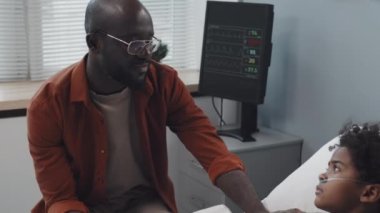 Tilting down and up of young African American man sitting by bed in hospital ward at daytime, talking to and holding hand of sick son with oxygen tube in nose