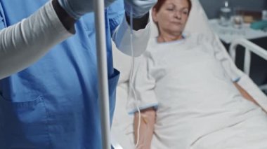 Tilting up of young Black male nurse wearing scrub suit, gloves and eyeglasses adjusting dropper of mature red-haired Caucasian woman who lying in hospital bed