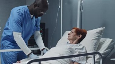 Önlük, eldiven ve gözlük takan genç siyahi erkek hemşirenin aşağı yukarı aşağı eğilmesi hastane yatağında yatan olgun kızıl saçlı beyaz kadına serum takması.