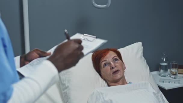 手術後に病院病棟に寝そべっていた赤髪の白人女性にインタビューし 医療記録に記録された青い頭皮の黒人男性看護師の肩越し — ストック動画