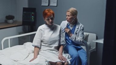 Medium long of blonde female doctor using stethoscope on mature Caucasian woman, sitting on hospital bed