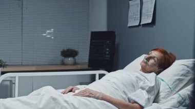 Medium long side view of older red-haired Caucasian woman with oxygen tube in nose lying in bed in room of hospital, looking away
