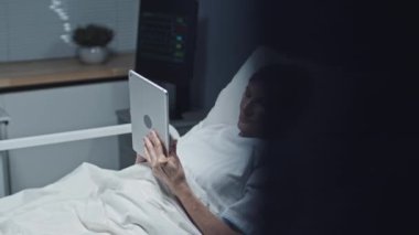Orta boylu, kısa saçlı, beyaz bir kadının klinikte yattığı tablet bilgisayar kullandığı ameliyattan sonra iyileşme sürecinde gülümsediği bir ortam.