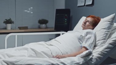 Medium long of older red-haired Caucasian woman sleeping in bed in clinic and smiling