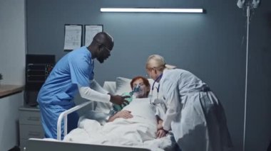 Hastane yatağında baygın yatan kızıl saçlı yaşlı kadın, sarışın beyaz kadın doktor ve hastaya koşan genç siyahi erkek hemşire, valf maskesi takıp onu hayata döndürüyor.