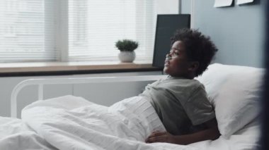 Close-up of African American boy with oxygen tube in nose sleeping in hospital bed at daytime, then waking up scared, mature Caucasian female doctor coming and comforting kid