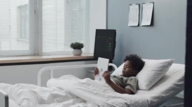 Full shot of Black boy with oxygen tube using tablet computer, lying in bed in hospital room at daytime