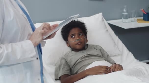 アフリカ系アメリカ人の男の子の中に酸素チューブがベッドに横たわっていて 病院の部屋に立っているタブレットコンピュータで医療ガウンを着た医師と話しています — ストック動画