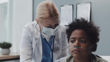 Medium of ill Black boy with oxygen tube sitting on bed in clinic room at daytime, Caucasian female pediatrician in medical mask and gown using stethoscope on his back, giving him instructions