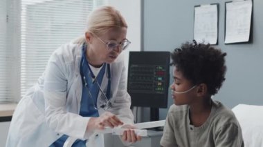 Medium long of Caucasian female pediatrician in medical gown standing in hospital ward at daytime, giving pill organizer to Black boy with oxygen tube in nose who lying in bed