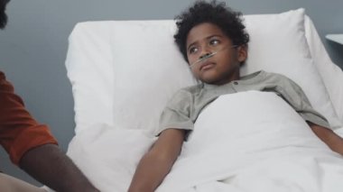 Tilting down and up of young Black man in eyeglasses sitting by bed in hospital ward at daytime, talking to and holding hand of unwell son with oxygen tube in nose