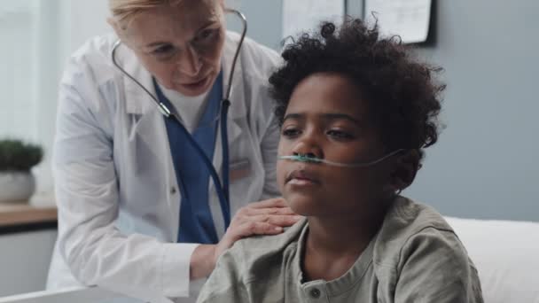 身穿卷曲的非洲裔美国男孩 头戴氧气筒 躺在病床上躺在病房里 白种人的金发女医生背着听诊器 与孩子们交谈 — 图库视频影像
