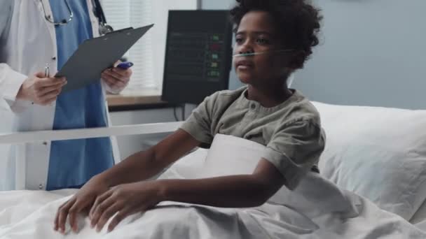 医師が来て 非接触温度計で彼の体の温度を測定するクロップ時に座って ベッドの上に横たわる鼻の中に酸素管を持つアフリカ系アメリカ人の少年のロックダウン — ストック動画