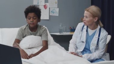 Medium of Black boy with oxygen tube in nose sitting up on bed with blond female Caucasian doctor in hospital room at daytime, video calling on portable computer