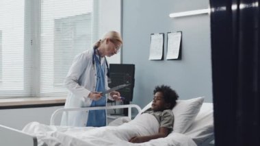 Locked down of Black boy with oxygen tube in nose lying in bed, then sitting up when female Caucasian doctor coming and measuring his body temperature with infrared thermometer