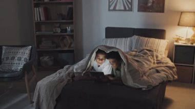 Tracking in of happy little African American girl and boy lying on stomach under blanket on bed at night, using tablet computer and talking