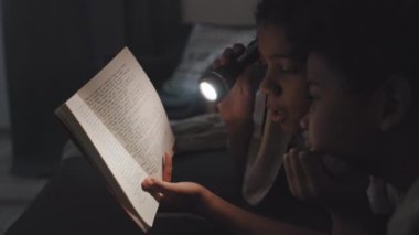 Side-view close-up of African American girl reading book using flashlight to little brother lying on stomachs under blanket on bed at night, smiling and talking