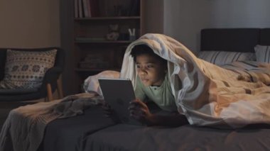 Medium long of cute Black boy lying on stomach under blanket in bed, using tablet computer at night, speaking