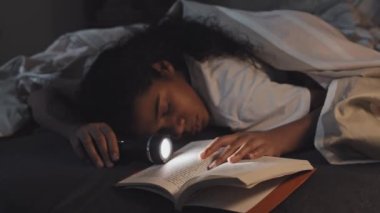 Low angle of African American tween sleeping on stomach under blanket in bed, lit flashlight and book in her hands