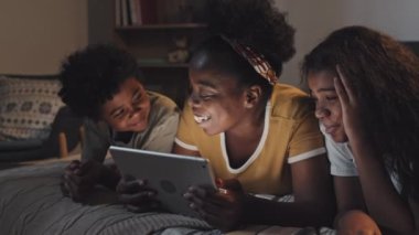Young African American woman lying on stomach in bed, using tablet computer at night, reading to daughter and son on her sides, smiling