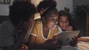 Chest-up of joyous young Black woman lying on stomach in bed, using tablet computer at night, reading out loud to cute little boy and girl on her sides, smiling