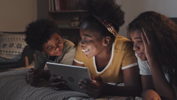 年轻的非洲裔美国妇女躺在床上 晚上用平板电脑给女儿和儿子读书 面带微笑 — 图库视频影像