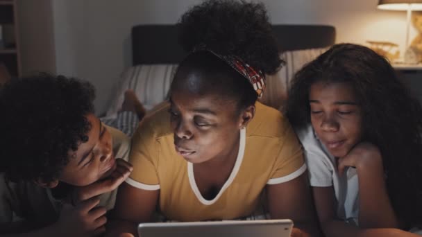 年轻的黑人妇女躺在床上 晚上用平板电脑 一边笑着一边大声念给可爱的侄女和侄子听 — 图库视频影像