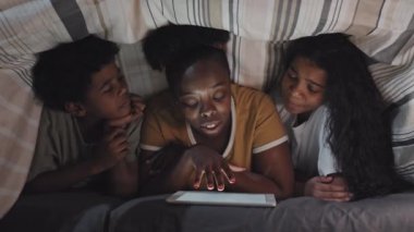 Chest-up of fun young Black mom lying on stomach covered with blanket, reading fairytale on tablet computer to adorable daughter and son on her sides, smiling