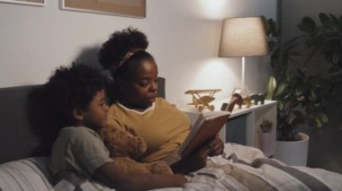 Medium of young African American mother reading book to cute nine-year-old son, lying in bed at night, boy holding toy bear