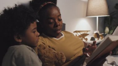 Medium close-up of young African American mother reading book to cute nine-year-old son, lying in bed at night, boy holding toy bear