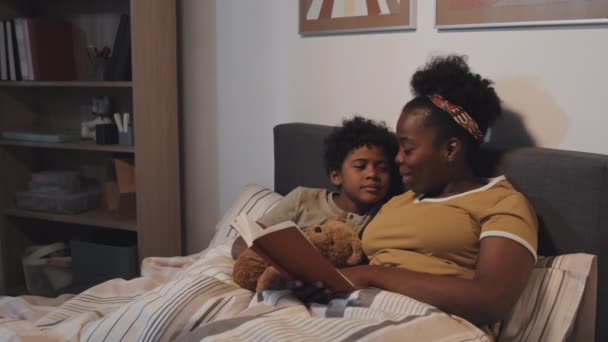 为可爱的九岁侄子读书的非洲裔美国姨妈的慢动作 晚上躺在床上 抱着玩具熊 — 图库视频影像
