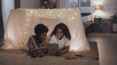 Chest-up of adorable African American brother and sister lying in magical blanket fort, using tablet computer at night, talking and smiling