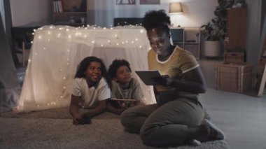 Full side view of young Black woman reading off tablet computer to cheerful little brother and sister who lying in magical blanket fort, smiling