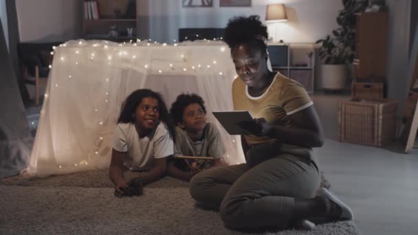 向躺在神奇的毛毯堡垒里笑着的快乐的弟弟和妹妹朗读平板电脑的年轻黑人妇女的全景 — 图库视频影像