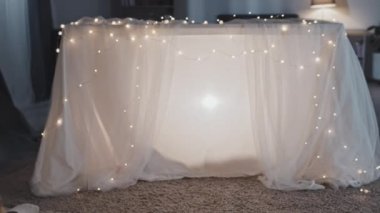 Tracking out rear of young African American woman and boy sitting on floor at home in dark, watching shadow puppets on white blanket with garland, clapping hands