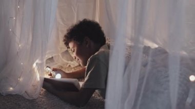 Medium side view of cute African American boy lying in dreamlike blanket fort, reading book using flashlight, smiling