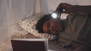 Medium close-up of joyous Black nine-year-old child lying on floor in handmade tent at home, reading book using flashlight at night, smiling