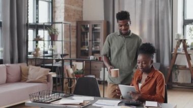 Locked-down of young African American woman sitting at desk at home in afternoon working remotely using tablet computer, her happy husband bringing coffee, talking