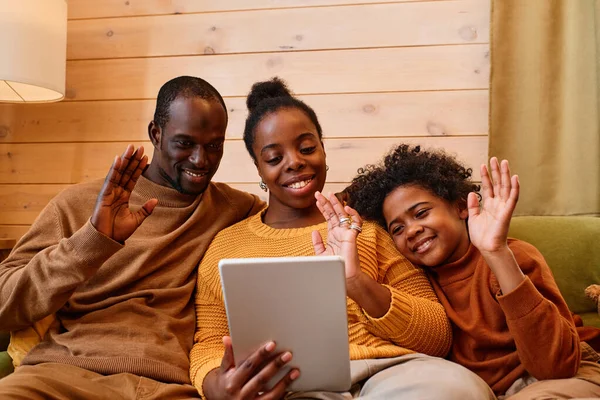 Happy young family with tablet communicating with their friends online while relaxing on sofa against wooden wall of their country house