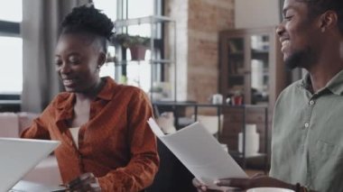 Tilting down of joyful African American couple sitting at desk in apartment in afternoon, smiling, high-fiving after having successful job done, getting up and leaving