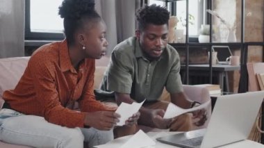 Medium long of young Black spouses sitting on couch in living room, doing paperwork, using portable computer and having conversation