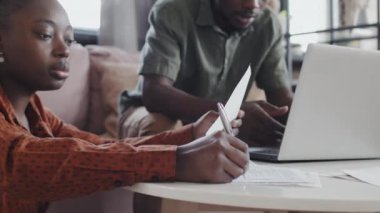 Medium close-up of young Black hetero couple sitting in living room, filling in tax returns, using portable computer and having conversation