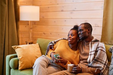 Happy young restful couple with cups of hot tea watching television in living room while relaxing on soft comfortable couch