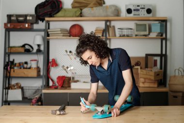 Youthful guy in blue shirt bending over blue skateboard on wooden table in garage of house while fitting wheels with handtool