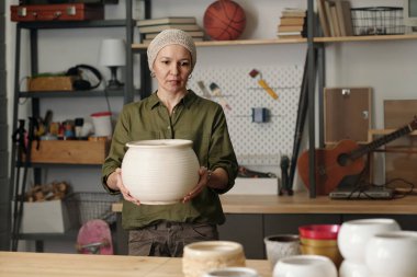 Contemporary retired female carrying large white clay vase or flowerpot for domestic plants while moving towards table in garage