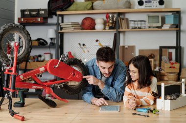 Young man pointing at tablet screen and wheel of bicycle while sitting by table in garage and explaining his little daughter how to repair it