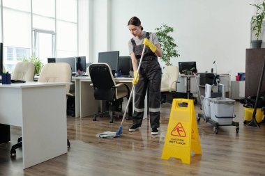 Young female cleaner in workwear using mop while cleaning floor in large modern openspace office with yellow plastic signboard in front