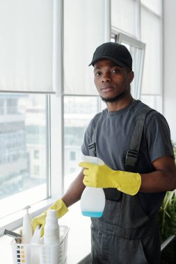 Contemporary young African American man with detergent in plastic bottle standing by sill with container while washing windows