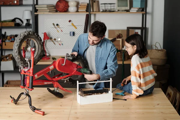 Young man using electric drill while repairing wheel of bicycle by wooden table while his daughter sitting near by and helping her father