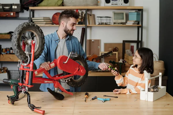 Cute little girl in striped pullover passing pliers to her father repairing bicycle by large wooden table in garage against household stuff